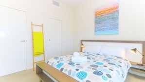 A bedroom with a bed, a bedside table with a lamp, a wooden ladder, and a painting on the wall. - Drift South 34 - Luxurious rooftop holiday apartment with lagoon pool views (Casuarina)