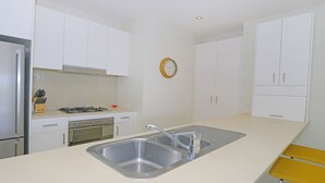 A modern kitchen with white cabinets, a stainless steel sink, a gas stove, and a yellow stool. - Drift South 34 - Luxurious rooftop holiday apartment with lagoon pool views (Casuarina)