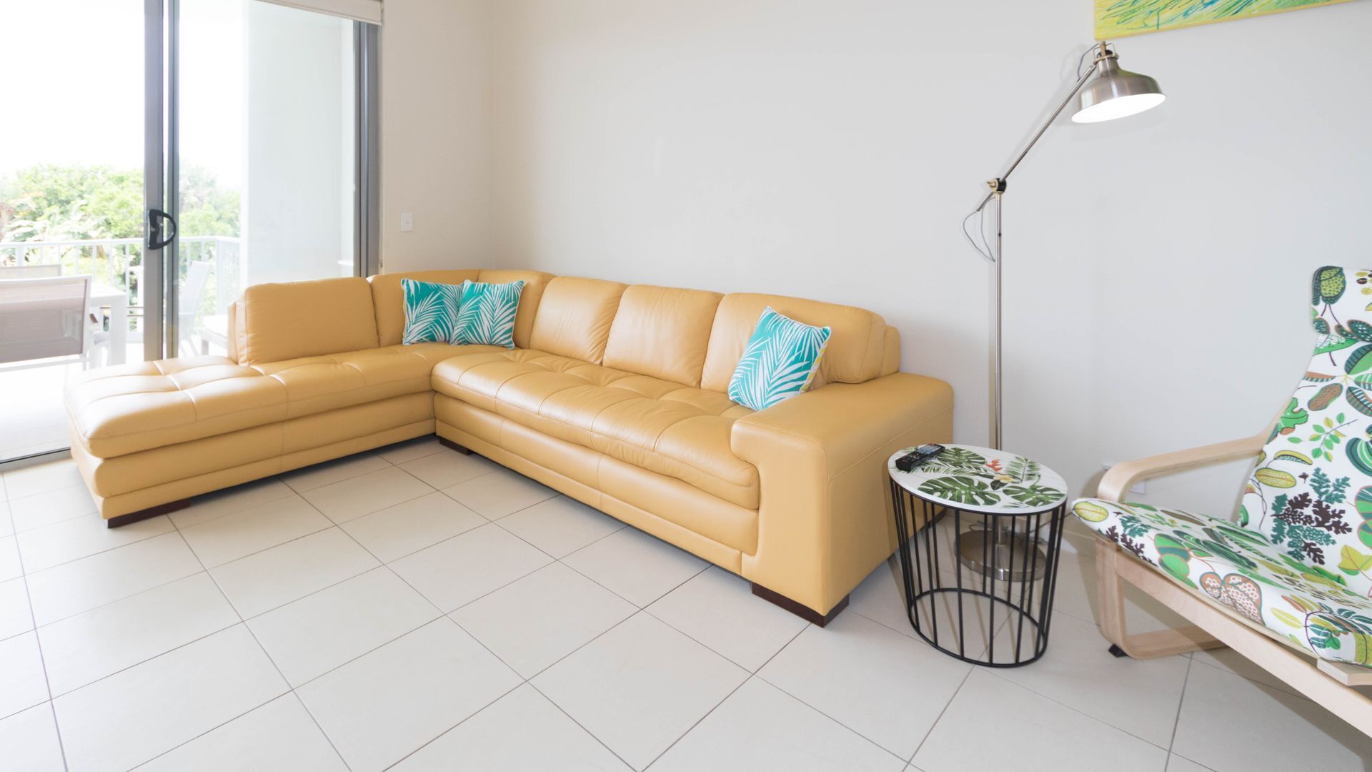 Ein Wohnzimmer mit einem beigefarbenen Ecksofa, einem kleinen runden Tisch, einer Stehlampe und einem gemusterten Sessel.