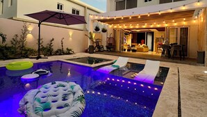 Un espace piscine extérieur moderne, doté d’un jacuzzi, de chaises longues et de guirlandes lumineuses.