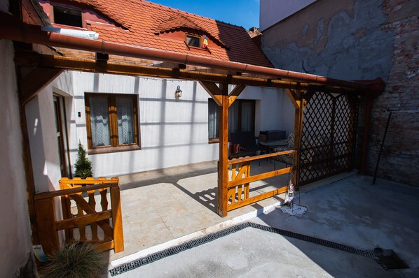 A courtyard with a wooden pergola, a small table, and a chair. There is a window with a curtain and a door with a metal gate.