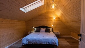 Ein gemütliches Schlafzimmer im Dachgeschoss mit schrägen Wänden, einem Bett mit gemustertem Überwurf, zwei Nachttischlampen und einem Oberlicht.
