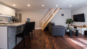 Eine moderne Küche mit integrierter Kochinsel, einem Essbereich mit schwarzem Stuhl, einem grauen Sofa und einer Holztreppe.