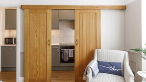 Une pièce dotée d’une porte coulissante en bois donnant sur une cuisine, d’un fauteuil à rayures avec un coussin et d’une table blanche.