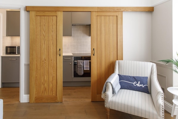 A room with a sliding wooden door leading to a kitchen, a striped armchair with a pillow, and a white table.