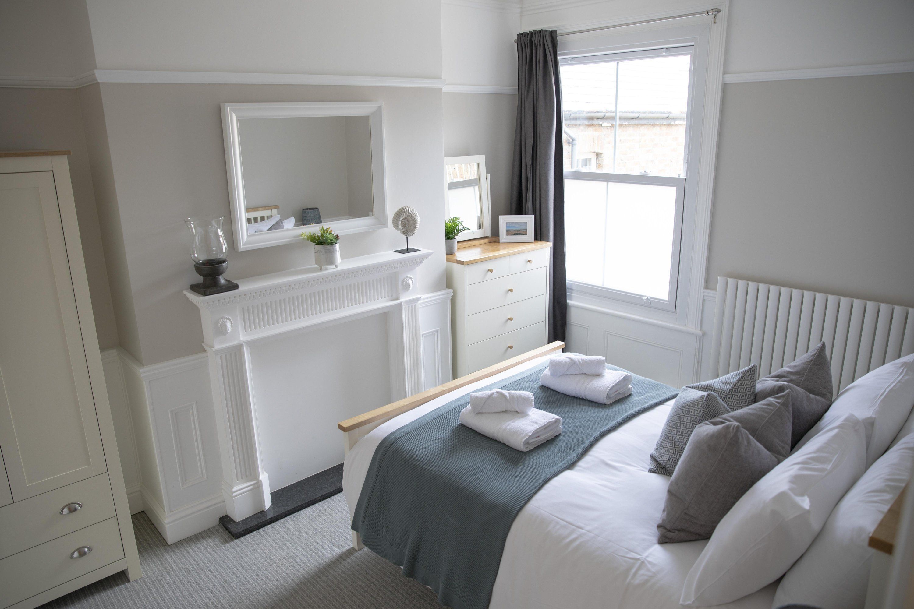 Un dormitorio ordenado con cama, cómoda, espejo y ventana con cortinas.