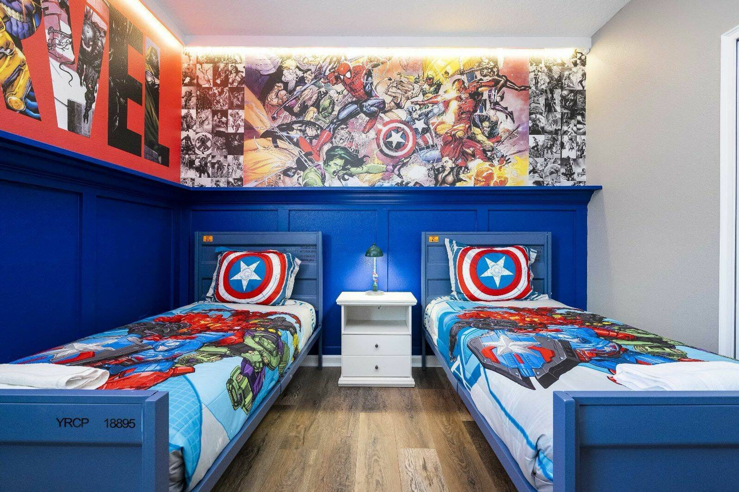 A room with two beds, each with a Captain America themed bedspread. The walls are blue, and there's a large comic book mural above the beds.