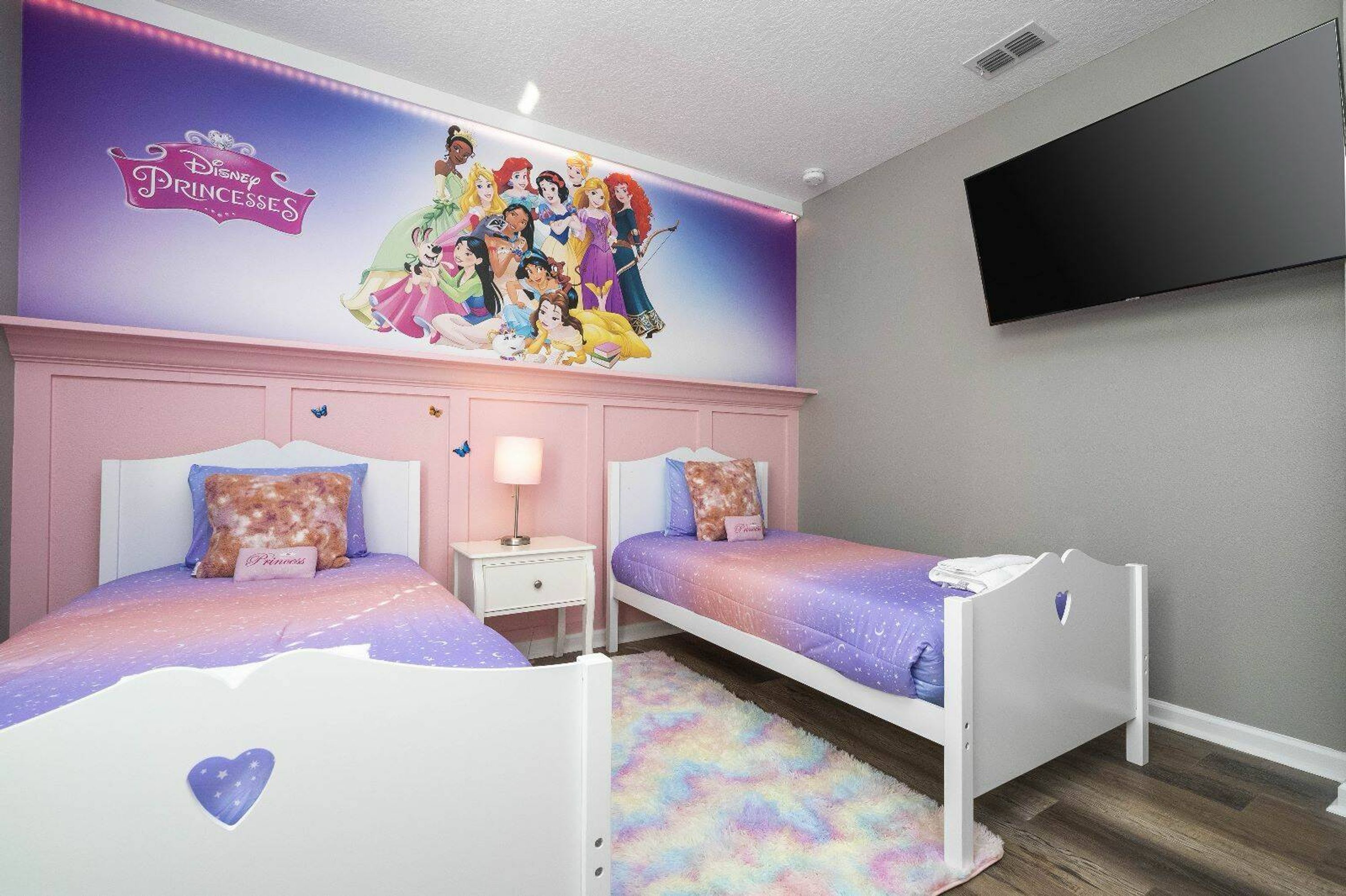 Camera per bambini con due letti, un affresco con le principesse Disney, una televisione a schermo piatto e un comodino.