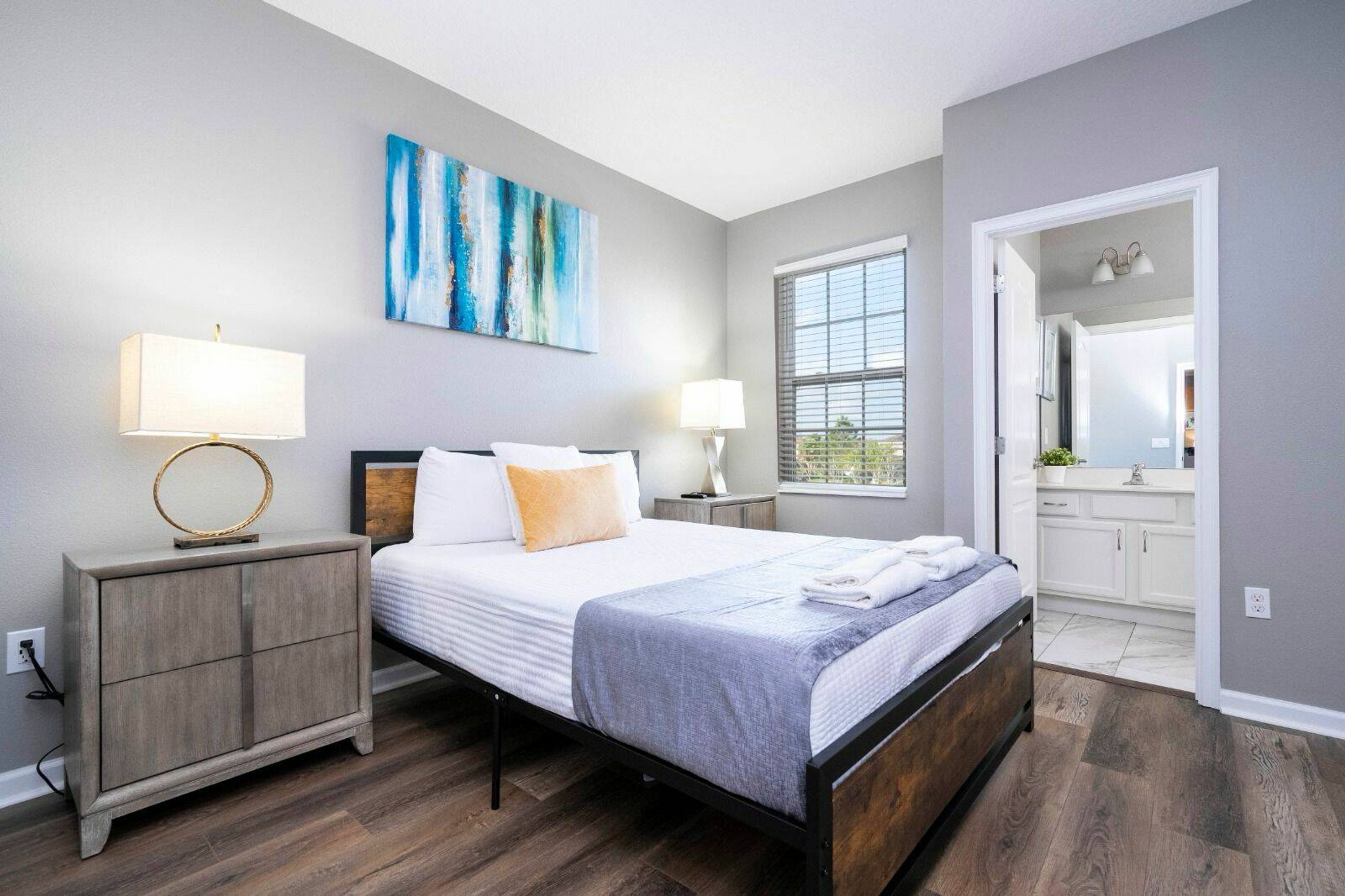 A modern bedroom with a bed, two bedside tables with lamps, a large window, and a bathroom visible through an open door.