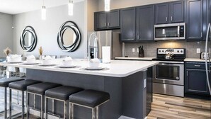 A modern kitchen with dark cabinets, stainless steel appliances, a central island with bar stools, and pendant lighting. - Solterra 12bd 11 5bat Sleeps24 Disney Pool Spa (Davenport)