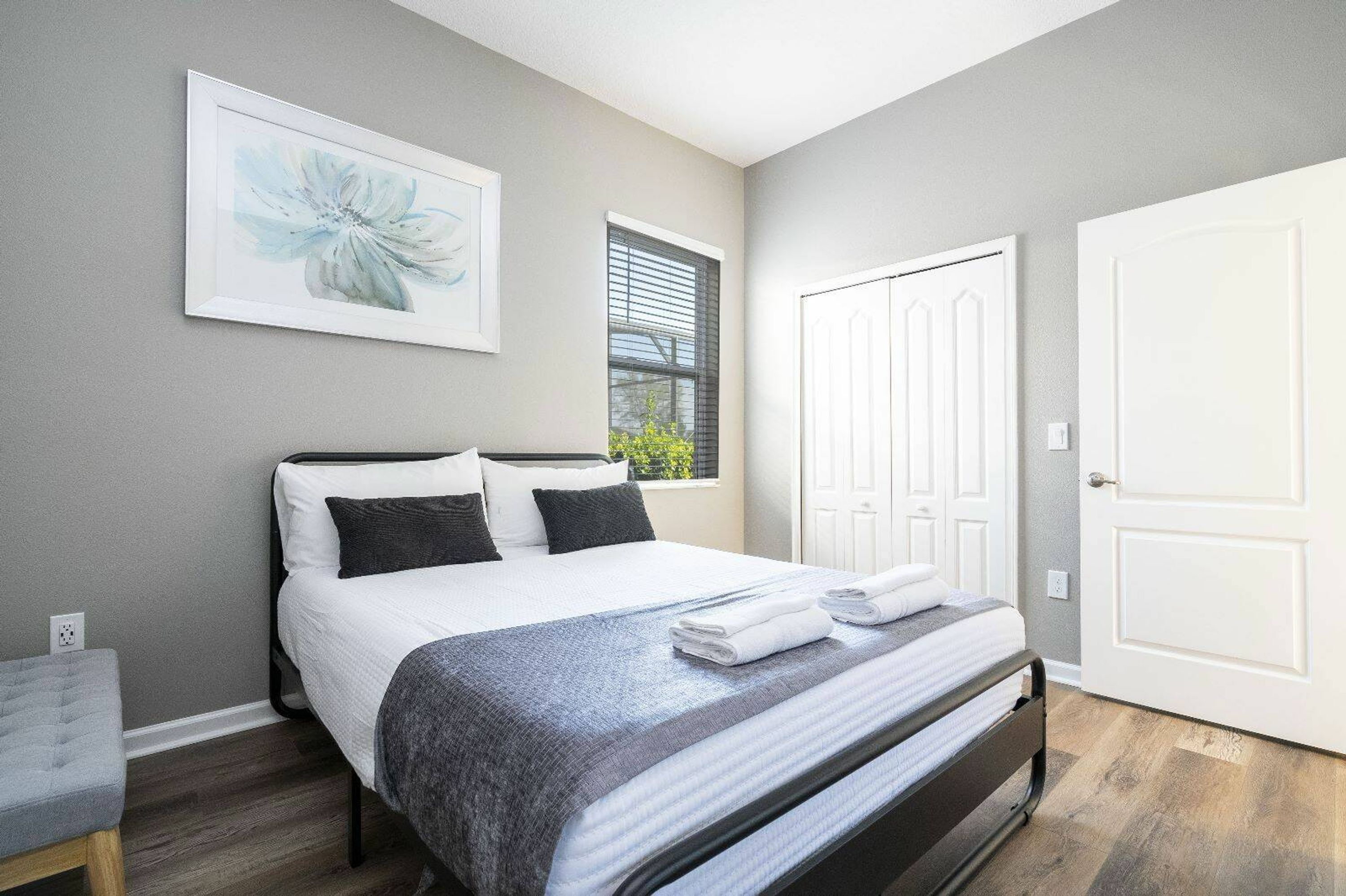 A neatly made bed with white and gray bedding, a framed floral artwork on the wall, a window with blinds, and a white door.