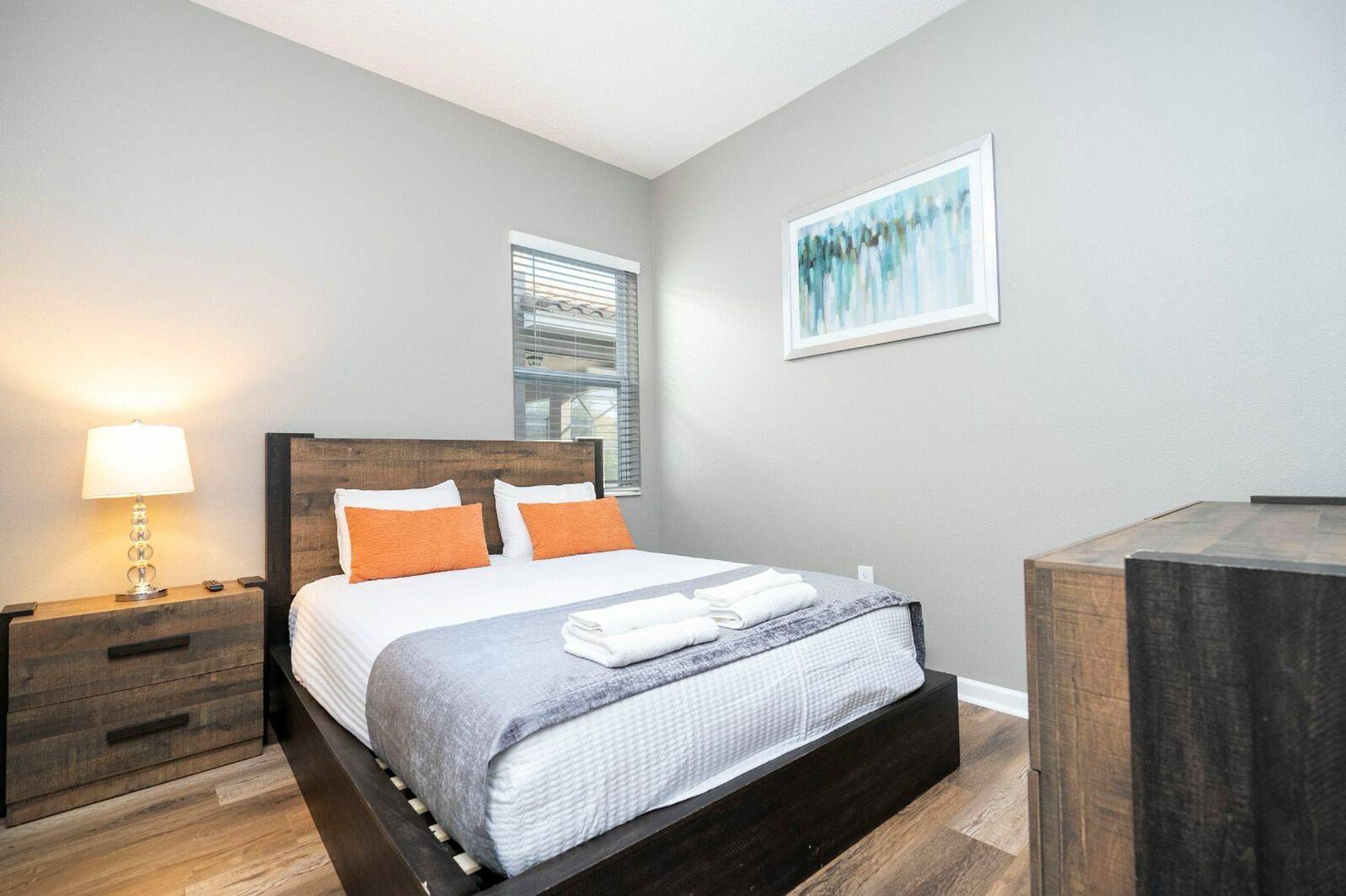 Un dormitorio ordenado con cama, mesitas de noche, lámpara y una obra enmarcada en la pared.