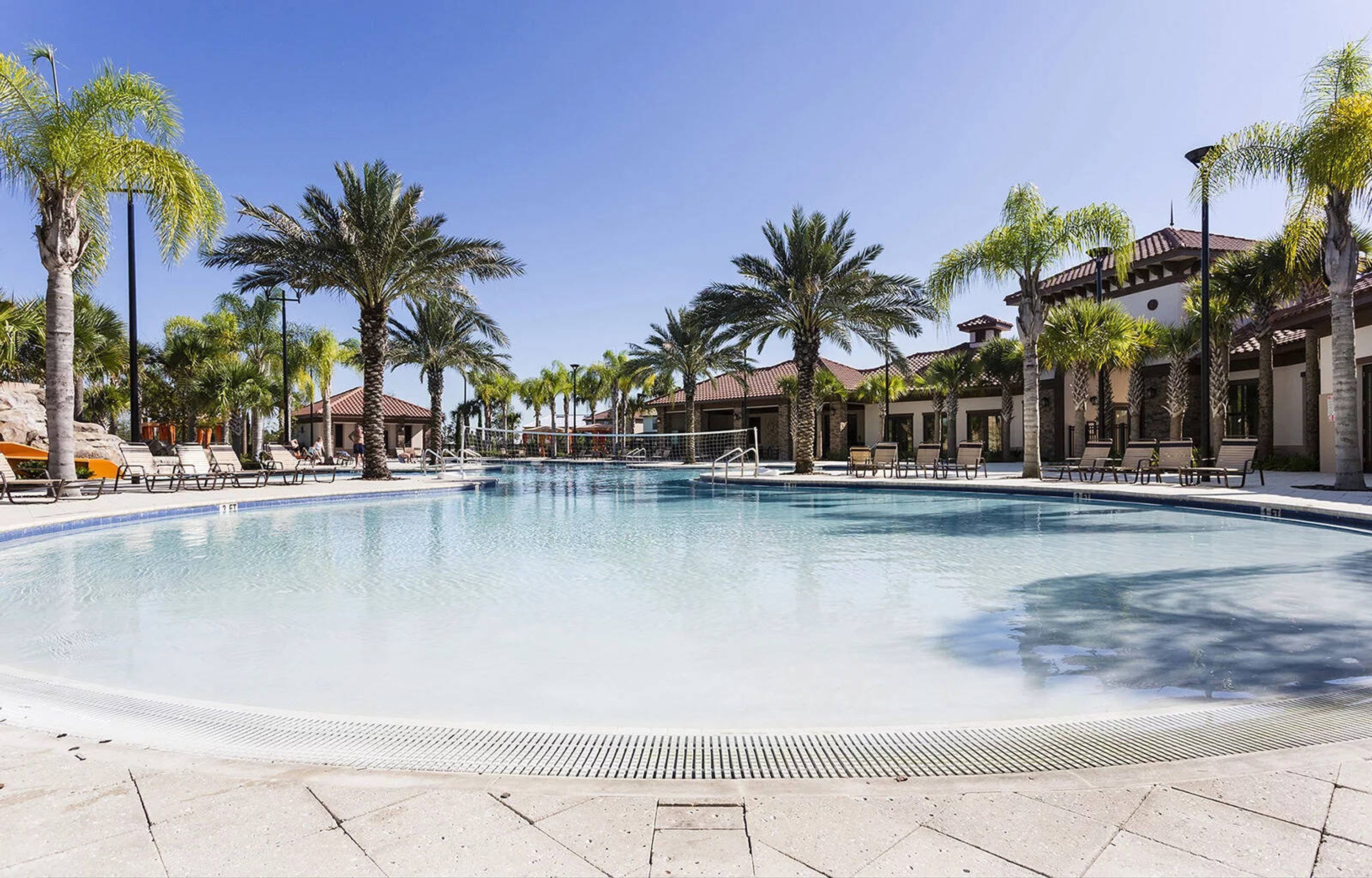 A large outdoor swimming pool surrounded by palm trees and lounge chairs, with buildings in the background.