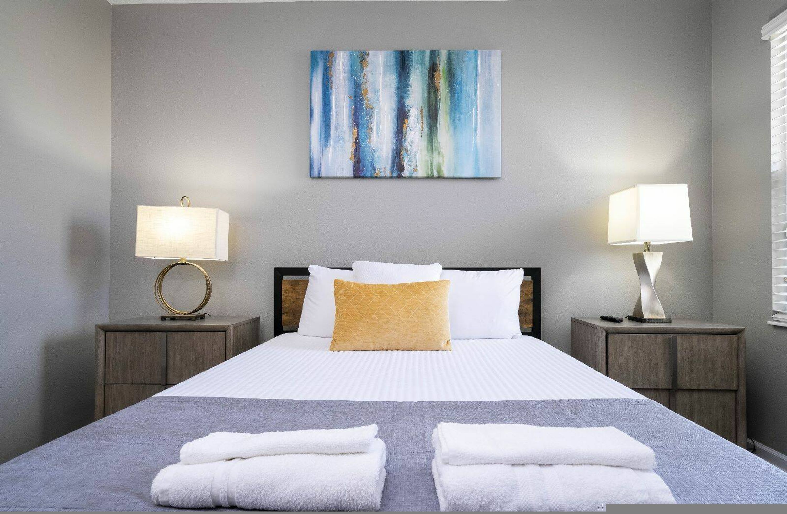 A neatly made bed with white linens and a single yellow pillow, flanked by two bedside tables with lamps, in a room with a gray wall and a blue abstract painting above the bed.