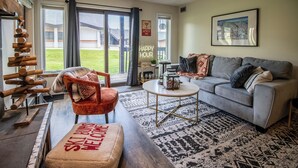 A spacious living room with a grey sofa, a red armchair, and a coffee table. A large window offers a view of a grassy yard.
