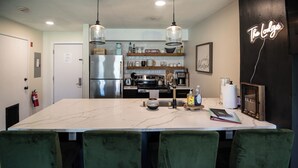 A modern kitchen with a large island, green upholstered chairs, stainless steel appliances, and a neon sign.