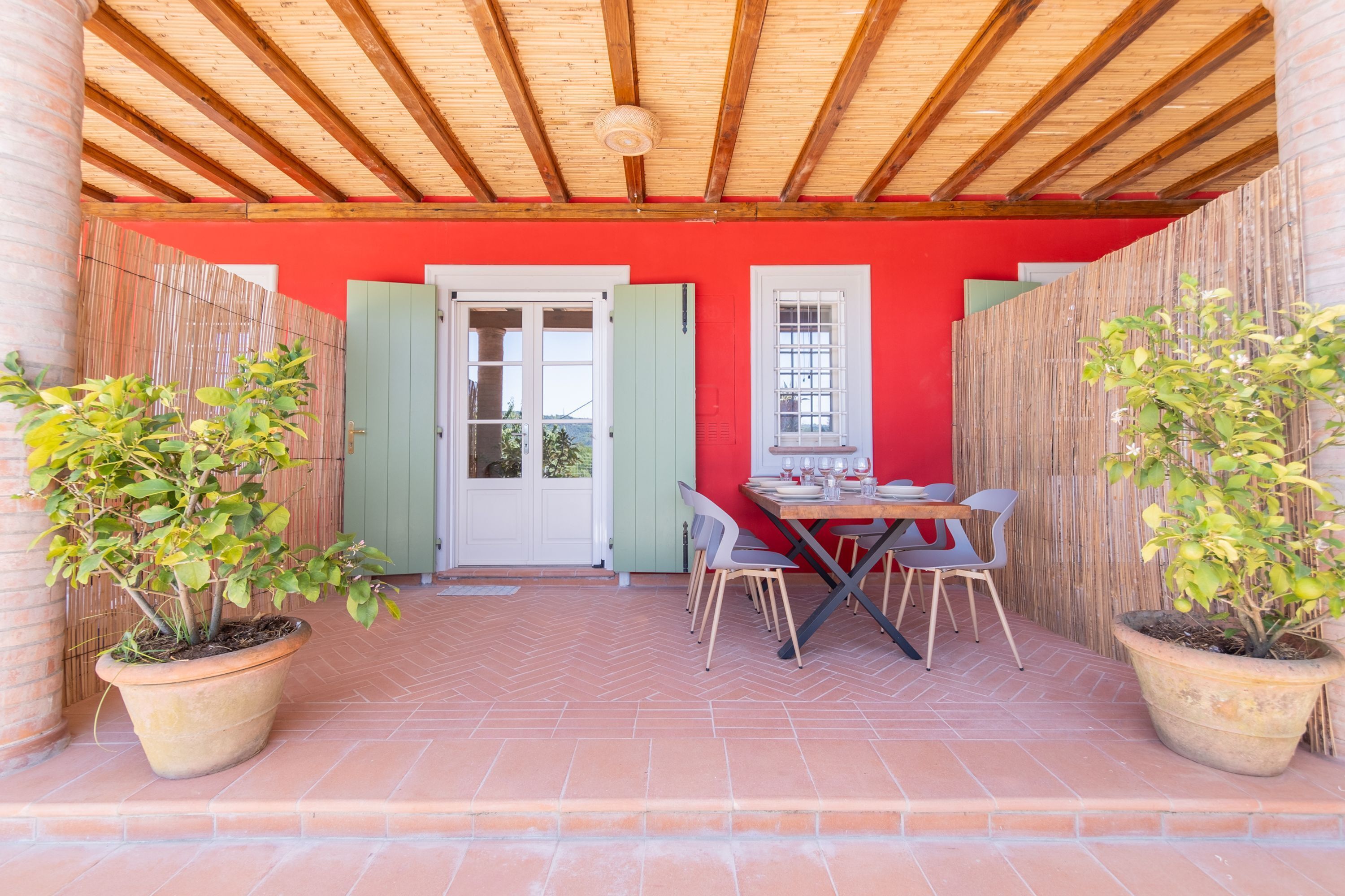 Een overdekt terras met houten plafond, een rode muur, en een tafel met stoelen. Er staan twee plantenpotten en een deur met groene luiken.