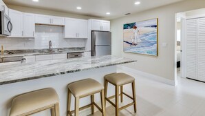 Una cocina moderna con armarios blancos, encimera de mármol, electrodomésticos de acero inoxidable y un cuadro con una escena de playa.