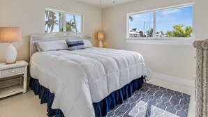 Un dormitorio con cama, dos lámparas de noche, un ventilador de techo y ventana con vistas a palmeras y cielo despejado.