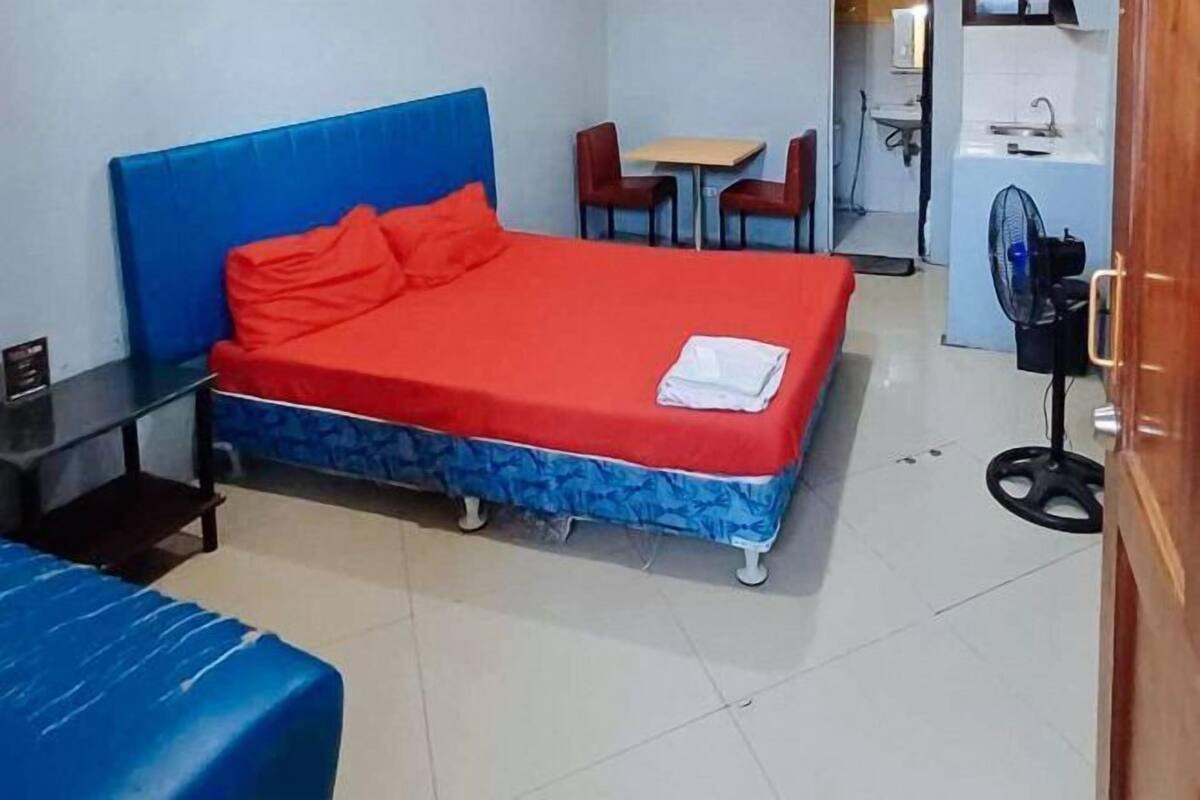 Una habitación con cama, una mesita, dos sillas, un escritorio y un ventilador.