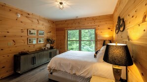 A wooden-paneled bedroom with a bed, bedside table, lamp, and a window with a view of trees. - The Dam House (Lake Lure)
