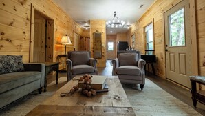 A log cabin interior with a wooden floor, two armchairs, a sofa, a coffee table with a book and fruit, a lamp, and a TV. - The Dam House (Lake Lure)