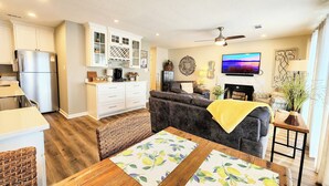 A modern living room with a sofa, a wooden dining table, and a kitchen area with white cabinets and a refrigerator. - Farr Shores Sunset Serenity (Hot Springs)