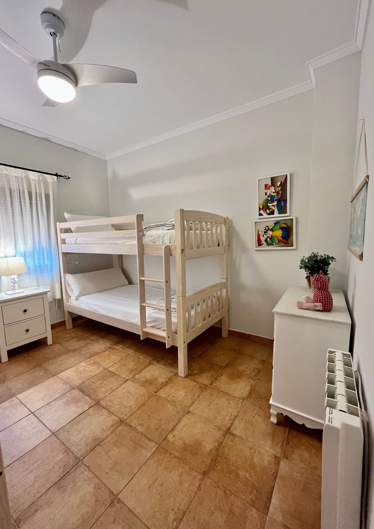A bunk bed room with a ceiling fan, a bedside table with a lamp, and a small white cabinet.