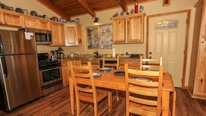 A kitchen with wooden cabinets, a stainless steel refrigerator, a wooden dining table with chairs, and a window with a view of trees. - Overlook Retreat- Awesome Views, Pets Welcome! (Big Bear City)