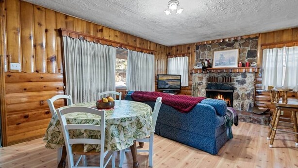 A log cabin interior with a stone fireplace, wooden walls, a dining table with chairs, and a blue sofa. - On Mountain Time-2bed 1bath+Loft (Big Bear City)