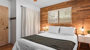 A bedroom with a wooden wall, a bed with white bedding, a window with blinds, a bedside table with a lamp, and a door with a coat rack. - Dutch Hideaway-3bed W/Gameroom (Big Bear City)