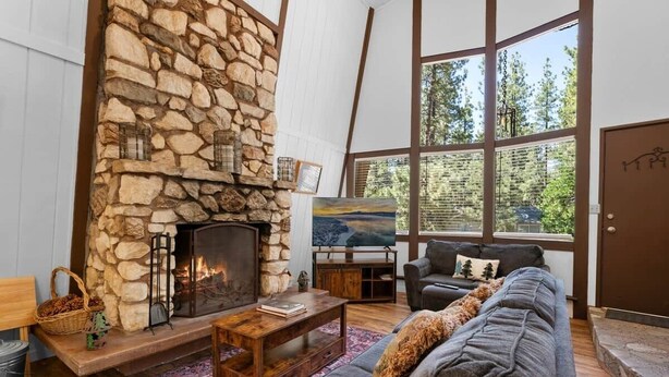 A cozy living room with a stone fireplace, a large window, and a comfortable seating area. - Sherwood Cabin-2bed+Loft (Big Bear City)