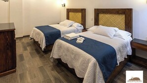 A hotel room with two beds, each with a wooden headboard, a blue bedspread, and a white lace cover. There is a wooden nightstand with a lamp on each side of the beds.