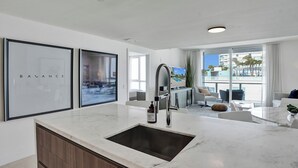 A modern kitchen with a marble countertop, a stainless steel sink, and a built-in oven. There are framed pictures on the wall and a view of the outside through large windows.
