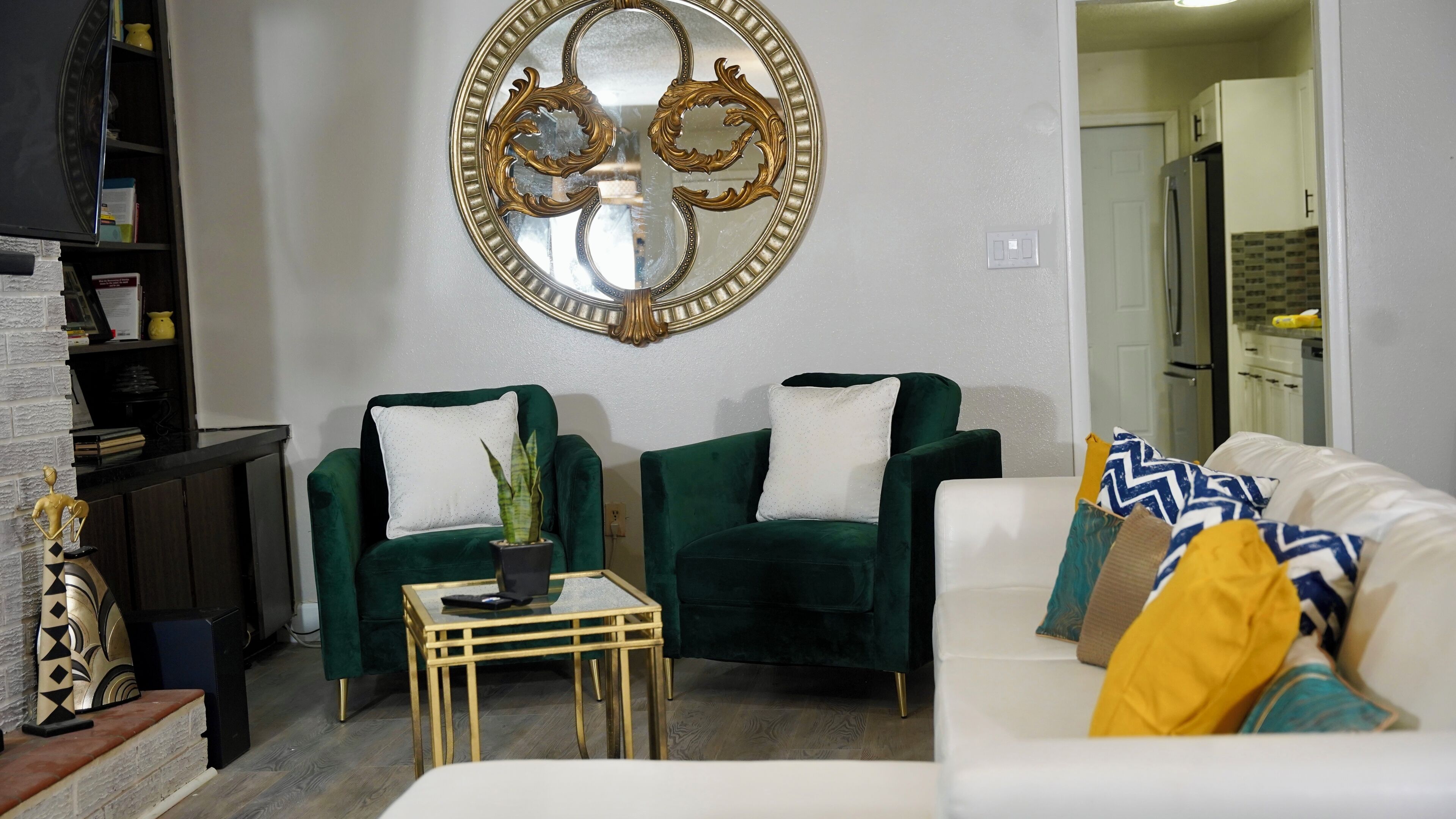 Un salon avec deux fauteuils verts, un canapé blanc et une petite table. Un miroir décoratif est accroché au mur, et on aperçoit une cuisine au fond.