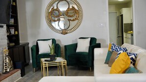 Un salón con dos sillones verdes, un sofá blanco y una pequeña mesa. Un espejo decorativo cuelga de la pared, y se ve una cocina al fondo.