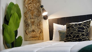 Una cama bien hecha con ropa blanca, cabecera negra y almohadones decorativos. Se observa una lámpara de pared, y una planta verde grande está colocada a la izquierda.