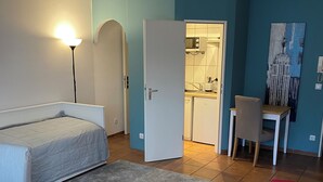Ett kompakt bostadsutrymme med en säng, ett skrivbord, en stol och ett litet kök.