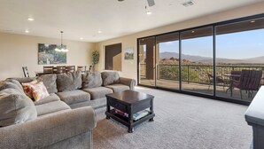 A spacious living room with a gray sectional sofa, a dark wooden coffee table, and a ceiling fan. Large glass doors lead to an outdoor patio with a view of mountains. - Luxury Desert Playground: Golf View + Resort Access (Hurricane)