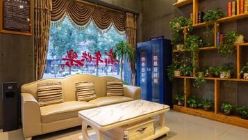 A hotel lobby with a sofa, a coffee table, a bookshelf with plants, and a blue information desk.
