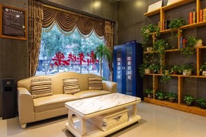 A hotel lobby with a sofa, a coffee table, a bookshelf with plants, and a blue information desk.