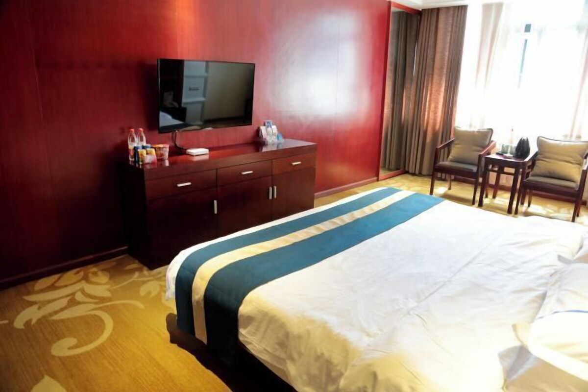 A hotel room with a large bed, a TV on the wall, a desk with a chair, and a window with curtains.