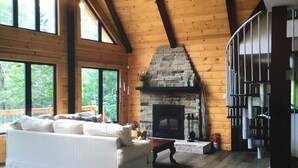 A spacious living room with a stone fireplace, wooden beams, large windows, and a cozy seating area. - Cottage life in Quebec  (Morin-Heights)