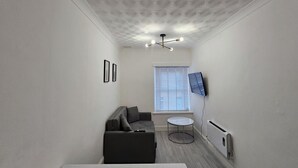 Un pasillo estrecho con techo estampado, un sofá gris, una mesita redonda pequeña y un televisor fijado a la pared.