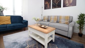 A living room with a blue sofa, a grey sofa, a wooden coffee table, and a large window with blinds. - 5 Bed House - Sleeps 16 - Parking - Wifi (Swansea)