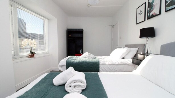 A hotel room with two beds, a window with blinds, a door, and a wall-mounted lamp. - 5 Bed House - Sleeps 16 - Parking - Wifi (Swansea)