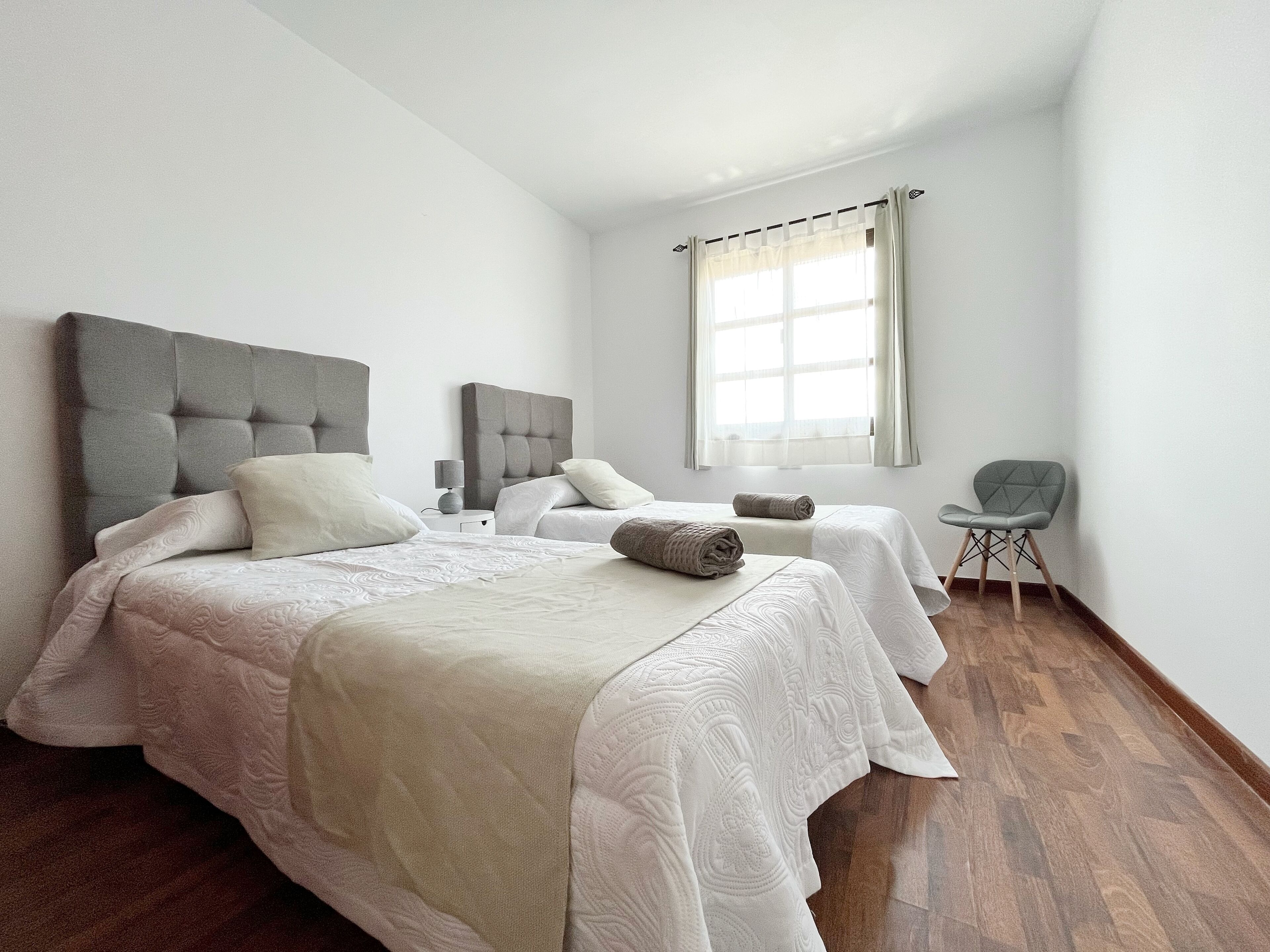 Une chambre moderne avec deux lits, une tête de lit rembourrée de couleur grise, une table de chevet avec une lampe et une fenêtre avec des rideaux.