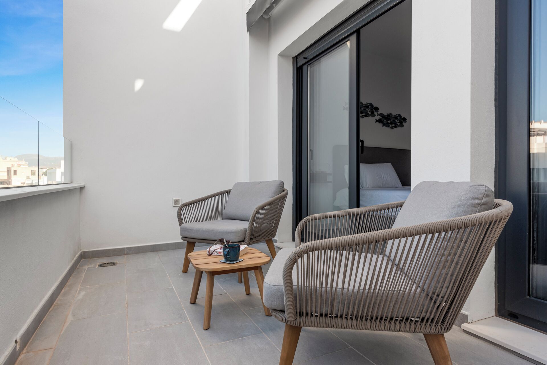 Un balcone con due poltrone in vimini, un tavolino di legno e un letto con materasso e cuscini bianchi.