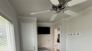 A bedroom with a bed, a ceiling fan, a TV, a bathroom, and a hallway leading to a kitchen. - Lake View Cabin, 20 miles from New Bern! (Maysville)