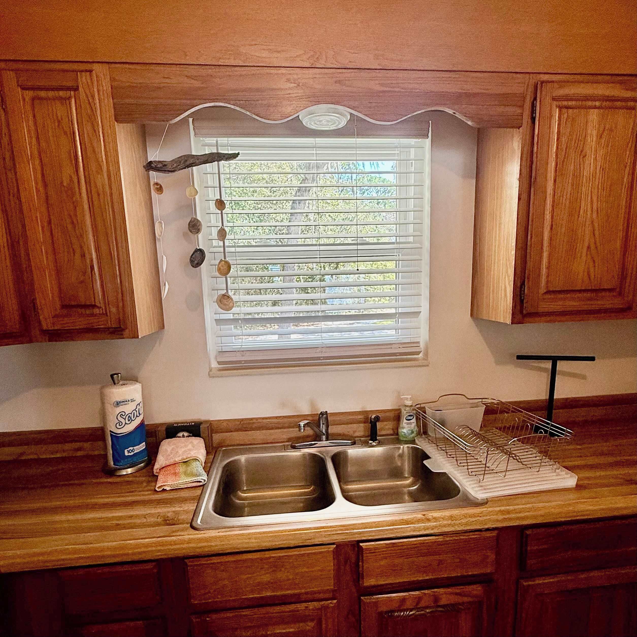 A kitchen with wooden cabinets, a double sink, a window with blinds, and a hanging rack.